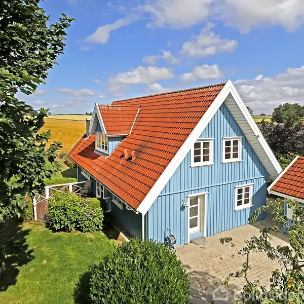 Blåt hus med rødt tag står i solskinsfyldt have, omringet af grønt græs og træer. Brolagt sti fører til hoveddøren. Bagved ses åbne marker og enkelte naboer.