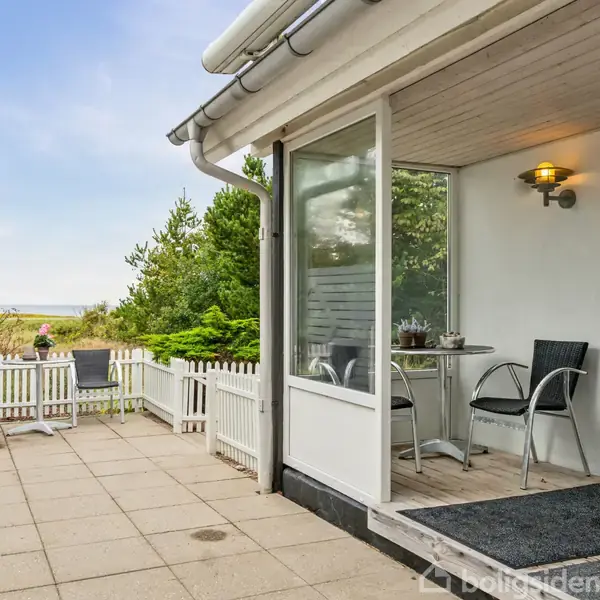 En terrasse med fliser har to stole og et bord under et halvtag. I baggrunden findes en hvid stakit, grøn beplantning og udsigt over et kystområde.