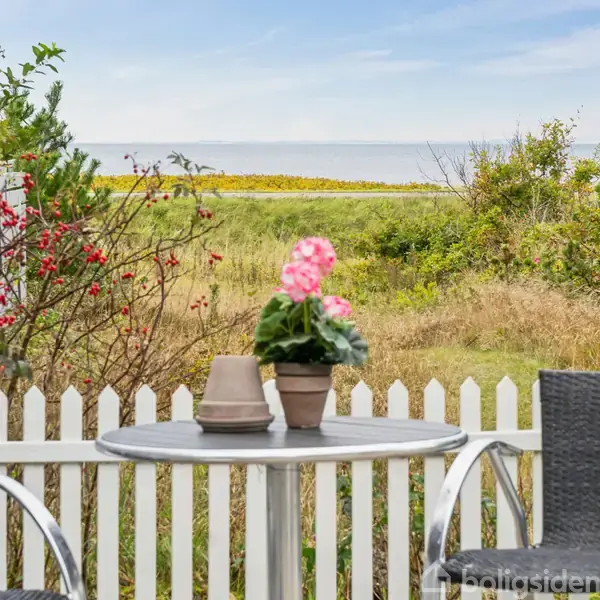 To sorte stole står omkring et rundt bord, som har en blomsterpotte og kop. I baggrunden er der en hvid stakit, buske og udsigt til havet.