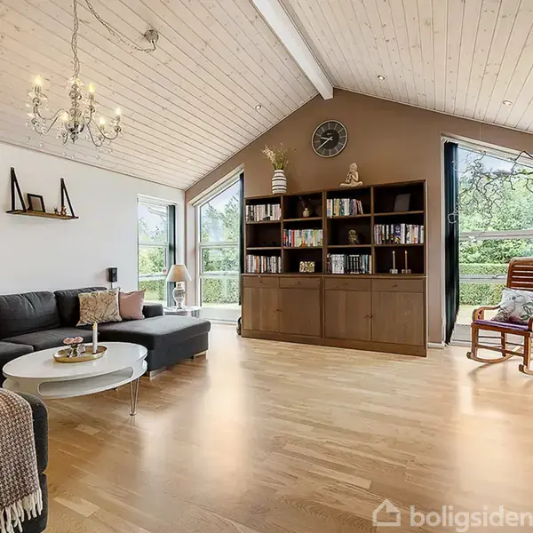Stue med mørk hjørnesofa, lavt sofabord og reol fyldt med bøger. En gyngestol står ved vinduet, og en lysekrone hænger fra loftet. Store vinduer slipper naturlys ind.