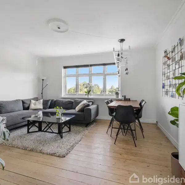 En grå sofa står i stuen op ad en hvid væg, flankeret af en lavalampe. Et sofabord og tæppe ligger på trægulvet. Ved vinduet står et spisebord med stole.