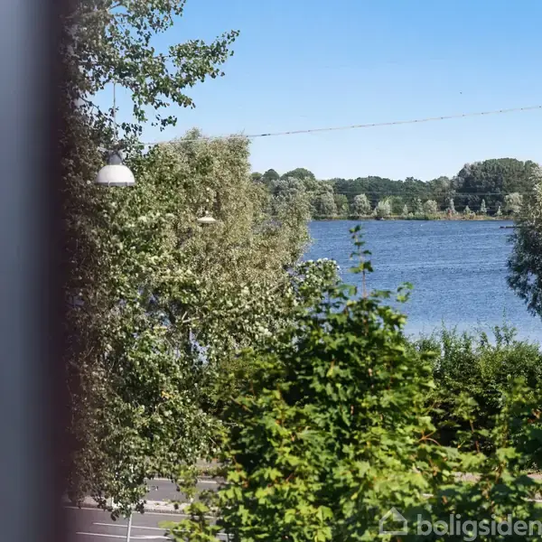 En sø ses gennem træer på en solrig dag. Grønne blade omgiver vandet, og himlen er klar. Et vejbelysningsarmatur hænger til venstre, og "Boligsiden" står nederst i højre hjørne.