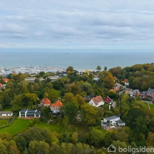 Kystby med hvide og røde tage ligger blandt træer. Baggrunden viser en marina med mange både, som strækker sig ud mod havet under en overskyet himmel.