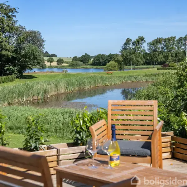 Flasker og vinglas står på et træbord på en terrasse med udsigt over en frodig have, en sø og en klar blå himmel i baggrunden.