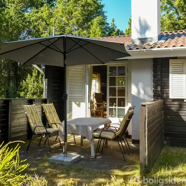 Et sort træhus med hvide skodder og en terrasse. Fire stole og et bord med parasol står på terrassen. Skoven omringer huset i solskinsvejr.