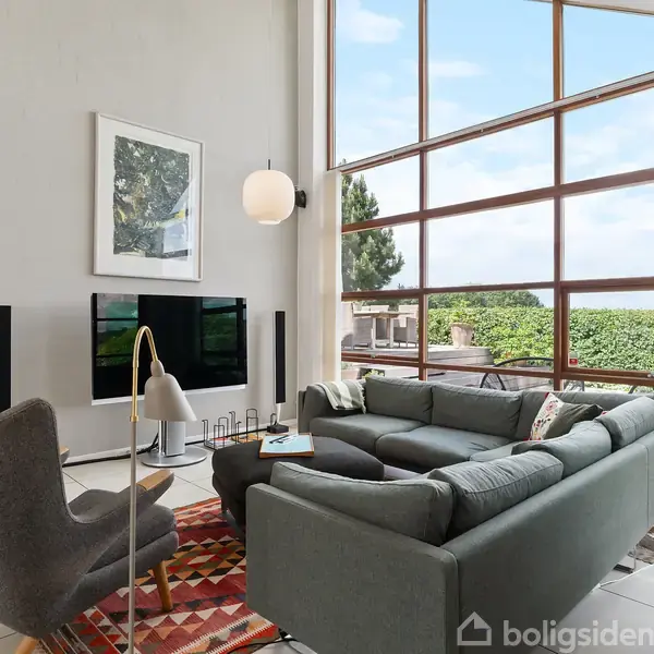 Grå sofa arrangeret i en stue med stor glasfacade, der giver udsigt til en have. En grå lænestol med gulvlampe står ved siden af et farverigt tæppe. Maleri på væggen.