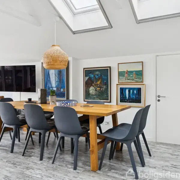 Et spisebord af træ er omgivet af seks blå stole. En loftslampe hænger over bordet. Væggen er pyntet med farverige malerier. Lys strømmer ind gennem loftvinduerne.