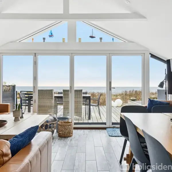 Stue med lædersofaer og spisebord åbner ud til en terrasse med havudsigt. Store vinduer slipper naturligt lys ind. Terrassemøbler udenfor. Ingen tekst bortset fra vandmærket "Boligsiden".