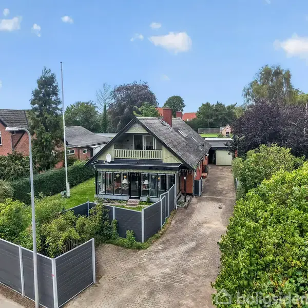 Et hus med en overetage, vinterhave og en lang indkørsel. Omgivet af hække og nabohuse i et villakvarter, ses et flagstang og parkeringsplads ved siden af huset.