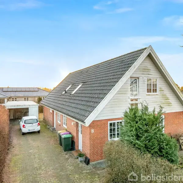 Et rødt murstenshus med gråt tag står i en beboelseskvarter. En hvid bil er parkeret i indkørslen ved siden af grønne affaldscontainere. Store buske omgiver huset.