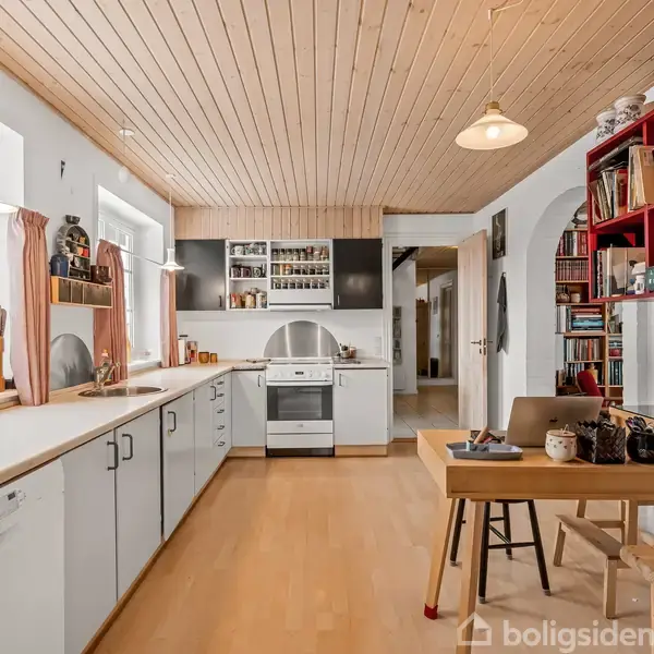 Et køkken med hvide skabe og lys træbordplade. En ovn står midt i rummet. Hylder med bøger og dekorationer ses på en rød reol til højre.