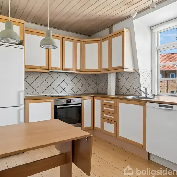 Køkken med lys træindretning og hvide skabe, køleskab og ovn til stede, bord i midten. Vindue mod en murstensbygning med rødt tag, træloft og ophængte lamper.