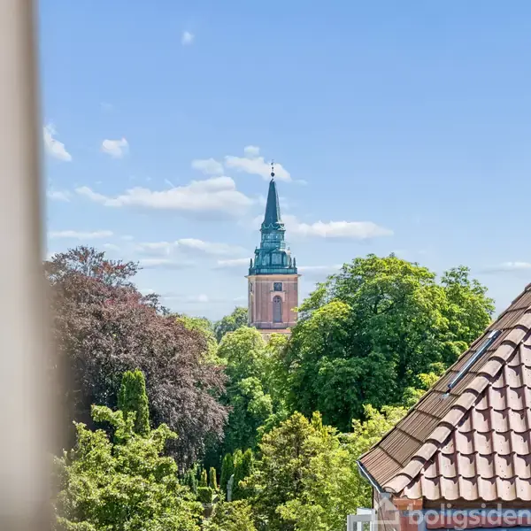Kirketårn rager op over grønne trækroner, med rødt tegltag til højre. Blå himmel med spredte skyer i baggrunden, skaber en idyllisk scene af natur og arkitektur.
