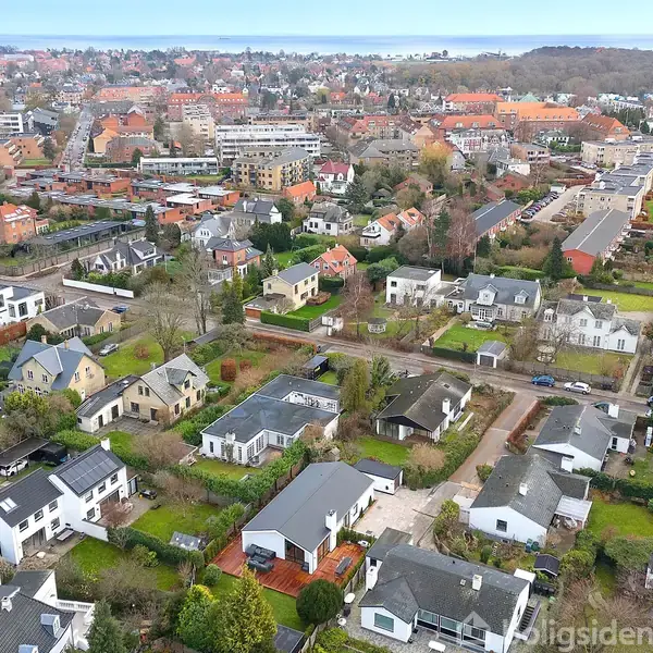 En gruppe huse står stille i en rolig forstad med grønne haver, omkranset af større byområder og med kysten synlig i baggrunden.