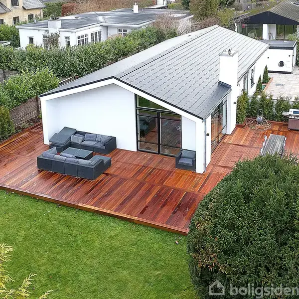 Moderne hus med hvid facade og sort tag ses fra oven. Træterrasse med grå udendørsmøbler omkranser huset. Grøn have omgiver området med buske og græs.