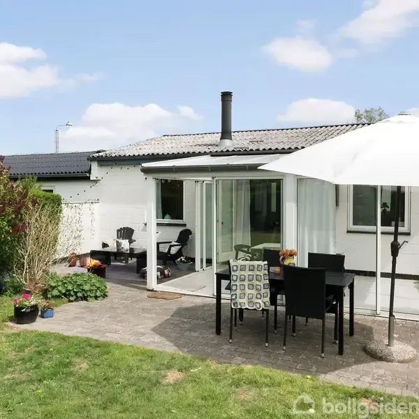 Hus med hvid facade, terrasse med havemøbler og parasol, set i en grøn have. Terrasseaktion viser siddemøbler under parasol. Omkringliggende planter skaber en indbydende atmosfære.