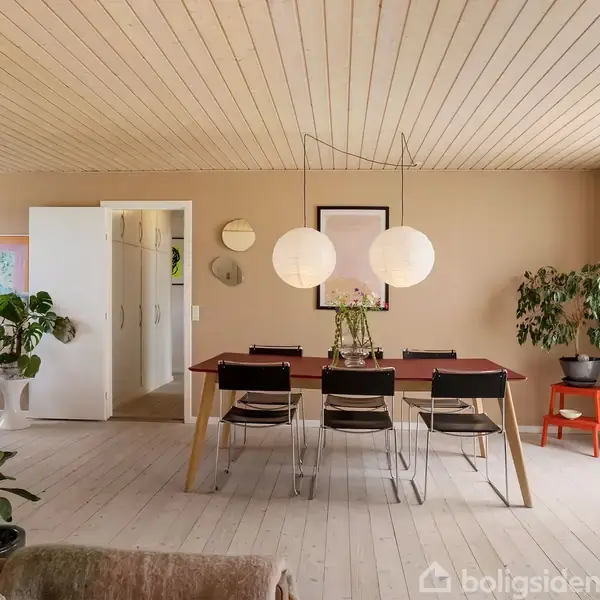 Et spisebord med stole står i en lys stue. To runde lamper hænger over bordet. Rummet har planter, kunst på væggene og en stor glasdør med udsigt til haven.