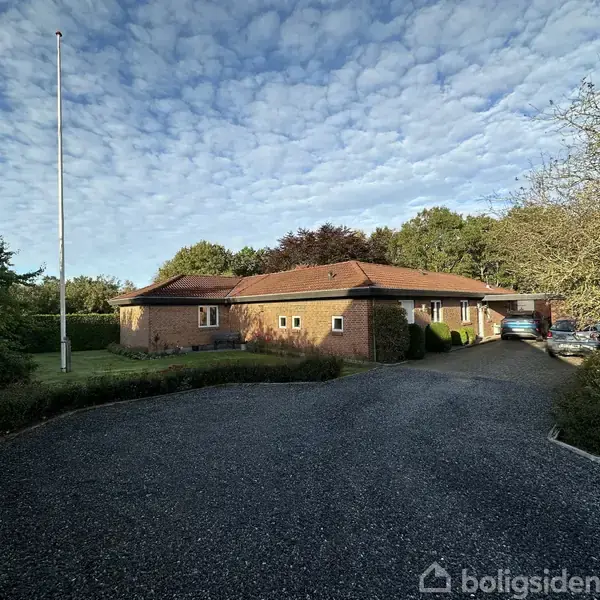 En fliseindkørsel leder til et murstenshus med rødt tag omgivet af en velplejet have og grønne træer. Biler er parkeret ved siden af huset.