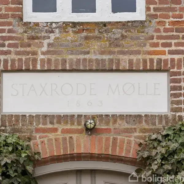 Skilt med teksten "Staxrode Mølle 1863" er indlejret i en murstensvæg over en dør. Murværket er omgivet af grønne klatreplanter.