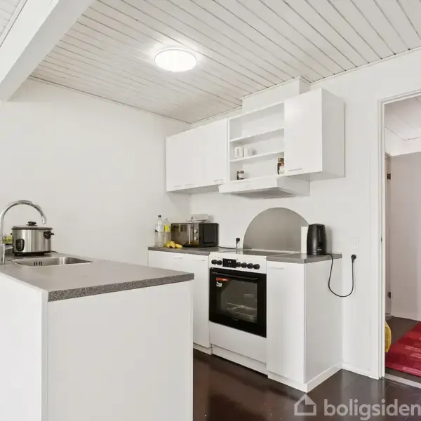 Køkken med en moderne ovn og kogeplade, omgivet af hvide skabe. På bordpladen står en kaffemaskine og en blender. Rummet har hvide vægge og loft, åbent mod en gang med rød løber.