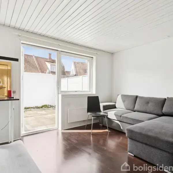 Grå hjørnesofa står i en lys stue med træloft og mørkt trægulv. En glashylde med to små figurer står ved en stor terrassedør, der åbner til udendørs gårdmiljø.