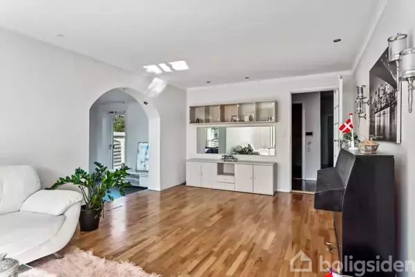 Hvid sofa står i stuen. Stueen med trægulv og spejlskænk mod væggen. Gennem bue er der udsigt til balkon. Lille dansk flag står på skænk.