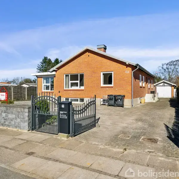 Rødt murstenshus står stille på en solrig dag. Omgivet af hæk og belægningssten. Indkørsel fører til hvid garage. Skilt til salg ved fortov.