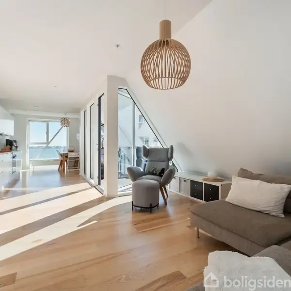 Stue med grå sofa og lænestol, oplyst af store vinduer. Et åbent køkken er i baggrunden. Moderne trælampe hænger fra loftet. Lys træparketgulv skaber en rummelig atmosfære.
