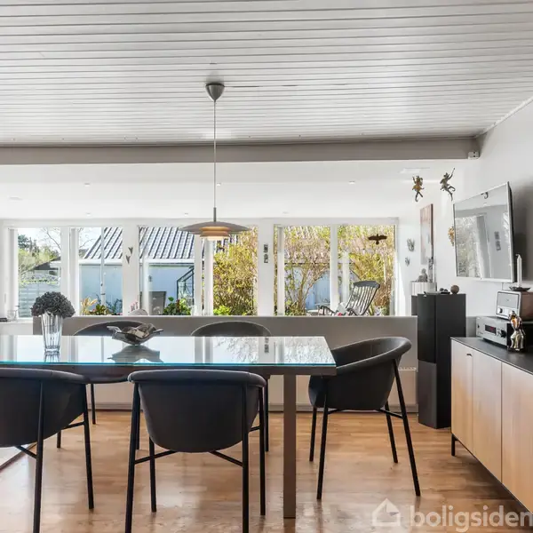 Et spisebord med sorte stole står i et lyst rum med store vinduer, som giver udsigt til en have. Et skænkbord med dekorationer er placeret til højre under et vægmonteret TV.