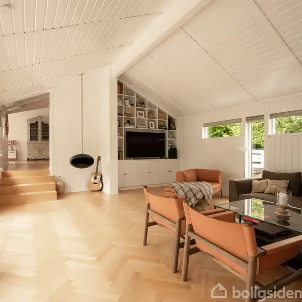 En stue med skrånende loft, store vinduer og lyse trægulve indeholder en hjørnesofa, to lænestole ved et glasbord, en guitar, og vægmonteret pejs. Trapper fører til et tilstødende rum.