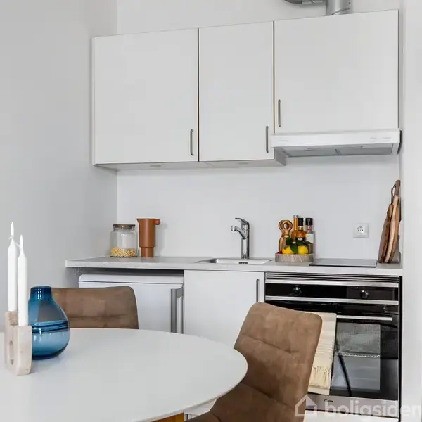 Et moderne køkken med en hvid bordplade og over- og underskabe. En rund spisebord står til venstre med to brune stole. Lys, skandinavisk indretning dominerer rummet.