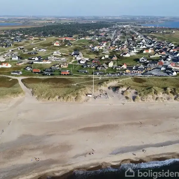 Strand med blødt sand strækker sig langs kysten; bagved ligger en by med spredte huse. Landskabet inkluderer græsmarker og havudsigt under en klar blå himmel.