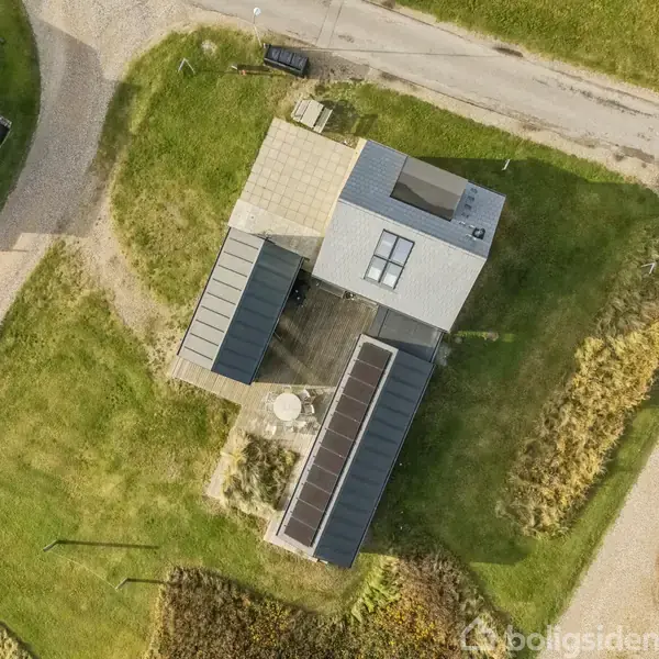 Et moderne hus set fra oven med solpaneler installeret på taget, omgivet af en grøn græsplæne og grusvej, beliggende i et beboelsesområde med flere andre bygninger i nærheden.