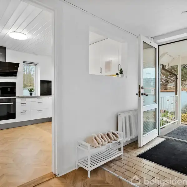 Entré med åbnet dør til forhaven, viser lyst køkken til venstre med hvide skabe og moderne apparater. Skohylde med sko ved en radiator. Udsigt til et grønt område udenfor.