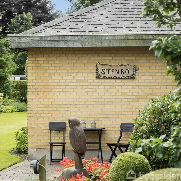 Et gult murstenshus med et skilt, hvor der står "STENBO". Foran huset står et udskåret dyreskulptur og et lille bord med to stole. Haven er velplejet med buske og græsplæne.