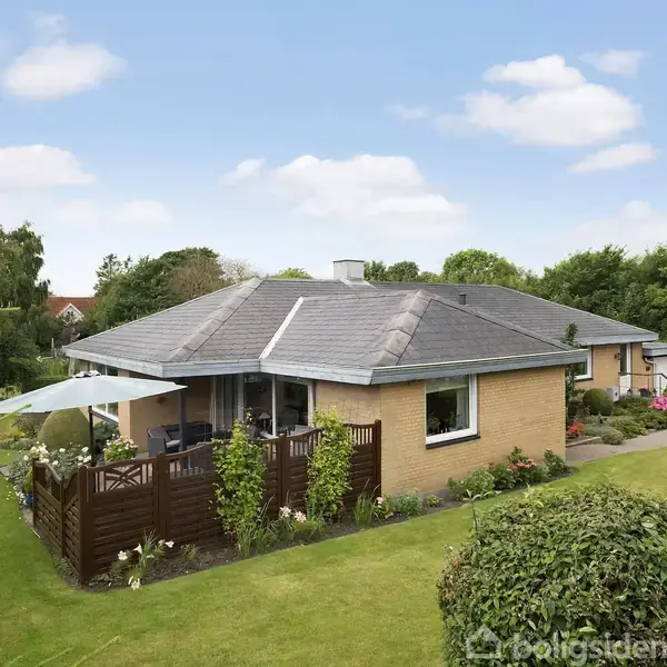 Et enfamiliehus i gule mursten med fladt tag står omgivet af en velplejet have. En terrasse med parasol er synlig foran, omgivet af frodig bevoksning og buske under en blå himmel.