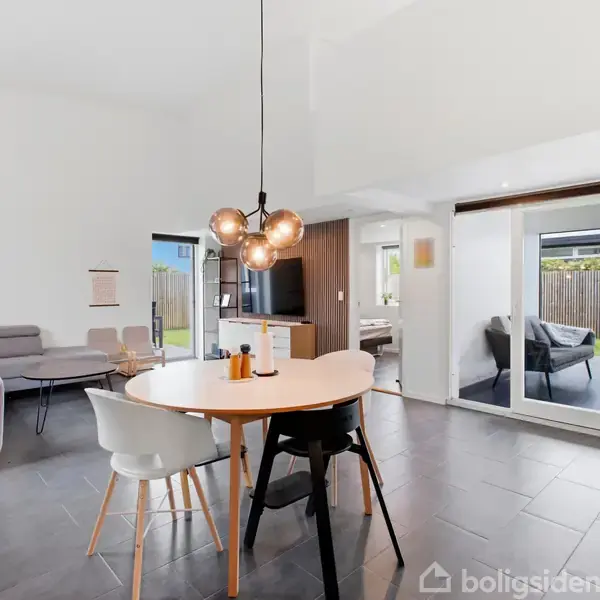 Et spisebord med tre stole står under en moderne lysekrone i en lys stue. Til venstre ses en grå sofa, og til højre store vinduer med udsigt til en have.