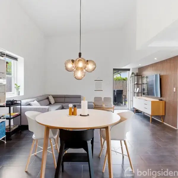 En rund spisebord med stole står i midten af en moderne stue. En sofa ses i baggrunden ved vinduet. Væggene er lyse, og der hænger en stilfuld loftslampe over bordet.
