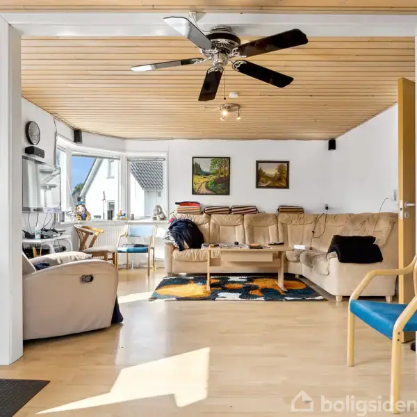 Stue med lysebrunt trægulv og loft, loftventilator i midten. Beige hjørnesofa og lænestol står langs væggen. Malerier hænger på væggen. Vinduer forsynet med persienner.