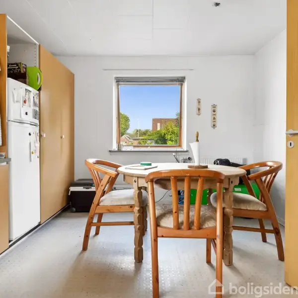 Et rundt bord med fire stole står i et lyst køkken. Køleskab og skabe til venstre, en udsigt til haven gennem et centralt placeret vindue i baggrunden.