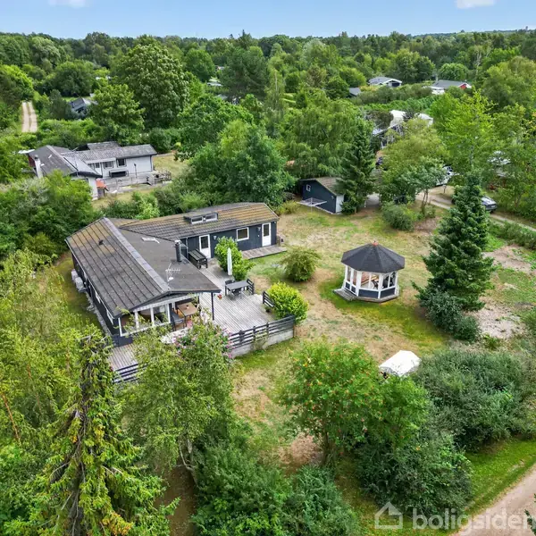 Hus med terrasse ligger i frodig have. I nærheden ses en lille, åbent pavillon. Omgivet af tæt skov og andre spredte bygninger i naturområde.