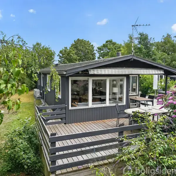 Sommerhus med træbeklædning står blandt frodige grønne træer; store vinduer viser ind i interiøret. Terrasse med bord og stole. Skyfri himmel i baggrunden.