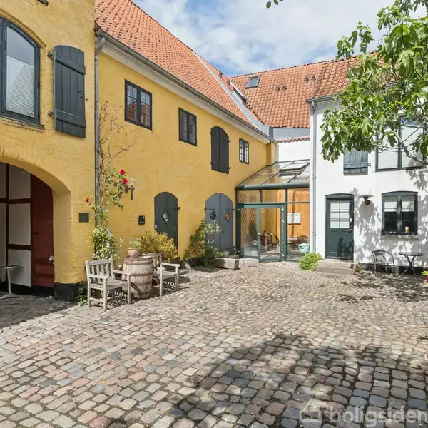 Gårdmiljø med brosten, omgivet af farverige bygninger med skodder. En gul bygning til venstre har en bueindgang og planter langs facaden. En hvid bygning til højre har vinduer.