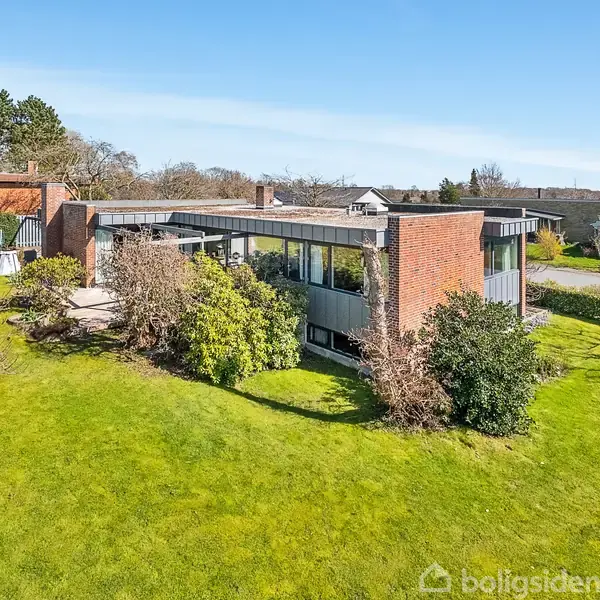 En moderne villa i ét plan med fladt tag, store vinduer og murstensvægge ligger i et åbent grønt område med en velplejet græsplæne og omgivende buske og træer.