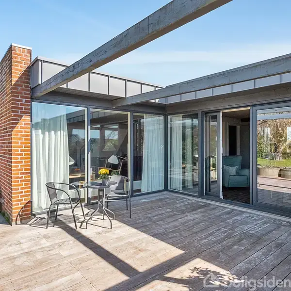 En terrasse med trædæk indeholder et lille rundt bord og to metalstole. Terrassedørene fører ind til en stue. Solen skinner, og der er beplantning rundt om.