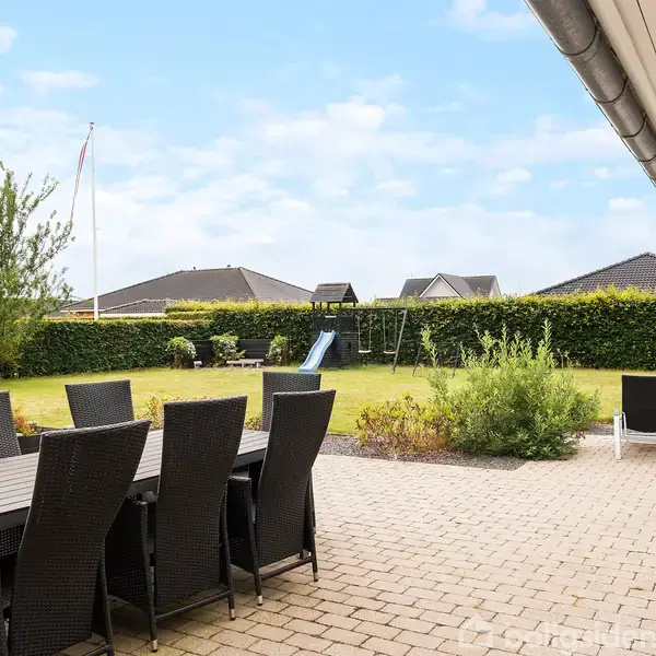 Terrassemøbler står på en flisebelagt terrasse med udsigt til en stor græsplæne med gyngestativ og rutsjebane. Hække og nabohuse omkranser haven under en blå himmel.