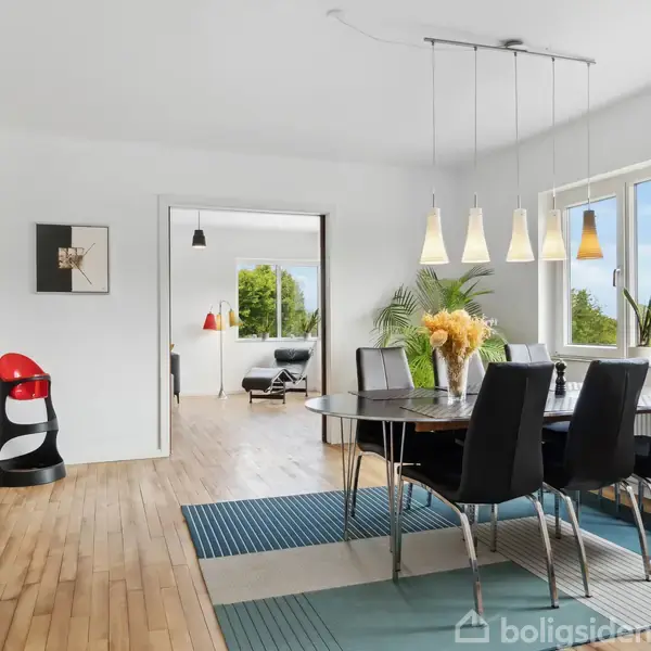 Et spisebord med sorte stole står i et lyst rum med store vinduer. I baggrunden ses en stue med planter og lyse vægge. Trægulv og moderne belysning præger indretningen.