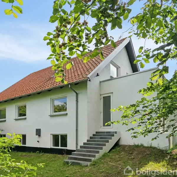 Hus med hvid facade og rødt tag står på græsplæne, omgivet af træer. Trapper fører til indgangsdør. Klar blå himmel ovenfor.