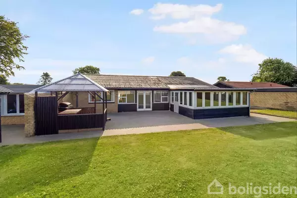 Hus med fladt tag og stor terrasse i en grøn have. Til højre er der en overdækket veranda med mange vinduer. I baggrunden ses et klart, solrigt himmelstræk.
