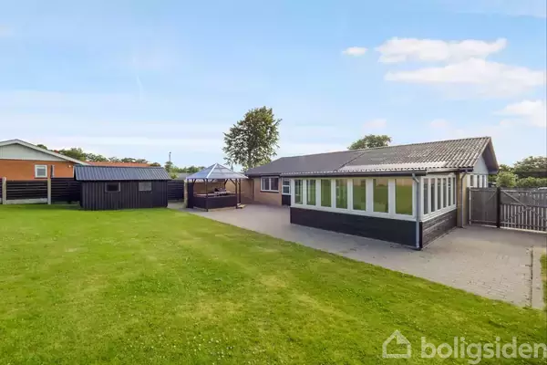 Hus med stor udestue står på flisebelagt terrasse. Græsplæne strækker sig omkring huset, med en træpavillon og et skur i baggrunden under en klar blå himmel.